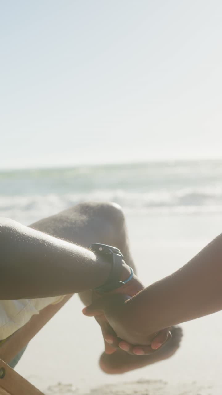 Vertical video of senior african american couple holding hands at beach, in slow motion