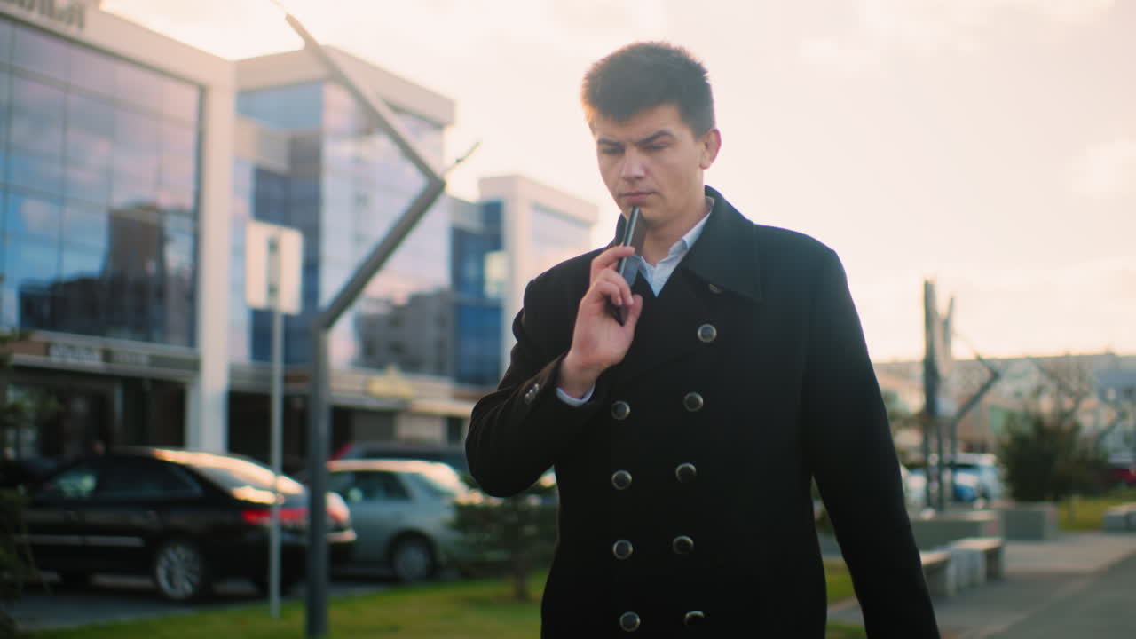 검은 코트를 입은 사업가가 전화 중에  에 전화를 들고, 현대적인 건물, 주차된 자동차, 그리고 배경의 초록색으로 도시 지역에서 밝은 빛 아래 자신감 있게 고 있습니다.
