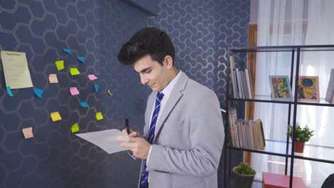 el hombre de negocios que hace el plan del día cuelga el papel en la pared y se sienta en su silla yendo a su escritorio.