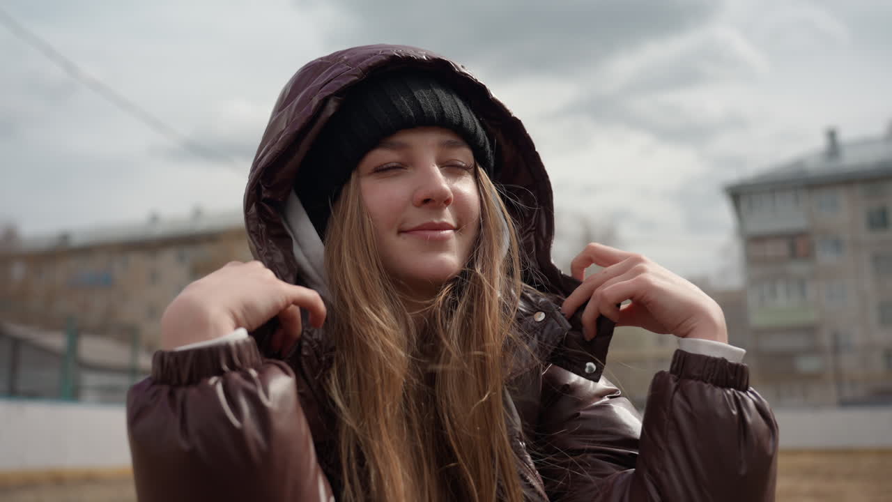 Mujer caucásica ajustándose la capucha al aire libre, con chaqueta acolchada y gorro de punto bajo un cielo nublado junto a edificios suburbanos, manos ajustando la capucha, sonrisa amable, brisa fría, retrato urbano espontáneo