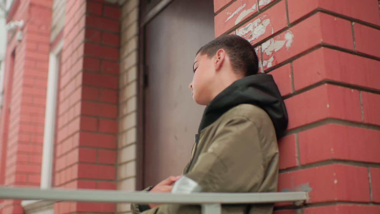 Hungry kid in hooded jacket leans on red brick wall near building entrance, gazing around thoughtfully with calm yet searching expression, capturing mood of quiet reflection