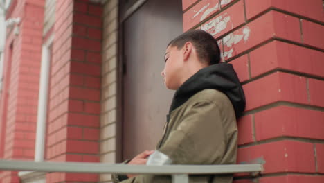 hungry kid in hooded jacket leans on red brick wall near building entrance, gazing around thoughtfully with calm yet searching expression, capturing mood of quiet reflection
