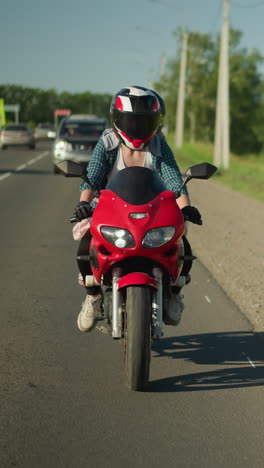 una mujer motociclista viaja lentamente por una carretera pavimentada con coches que se mueven en direcciones opuestas, postes eléctricos, árboles y edificios distantes son visibles en el fondo