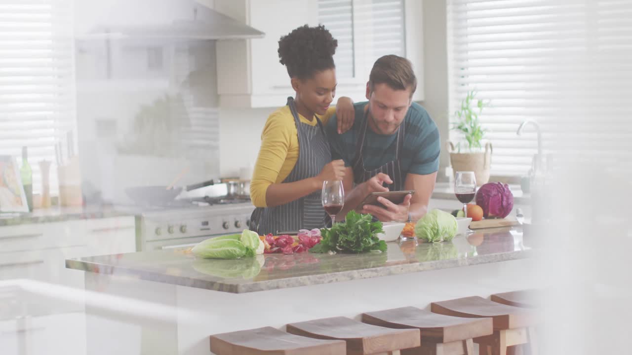 Video of happy diverse couple preparing meal, using tablet and drinking wine in kitchen