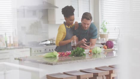 video of happy diverse couple preparing meal, using tablet and drinking wine in kitchen