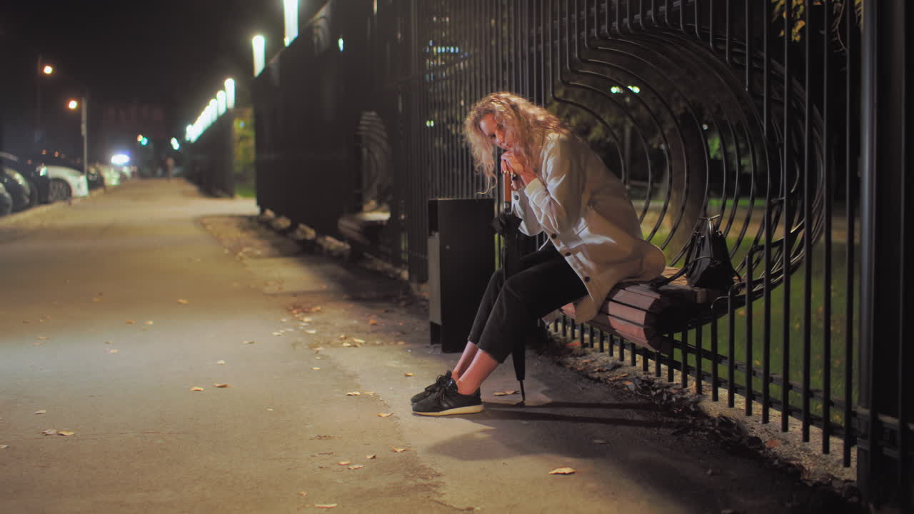Tired woman wearing white coat rests head on umbrella while seated on decorative iron fence bench with black bag beside her, surrounded by parked cars and illuminated by street lights