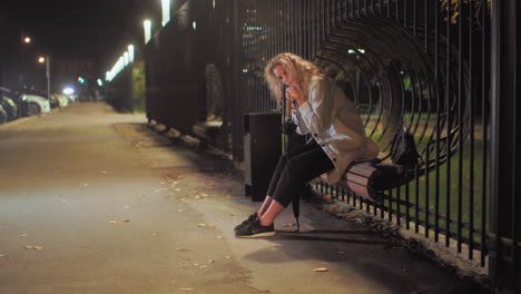 tired woman wearing white coat rests head on umbrella while seated on decorative iron fence bench with black bag beside her, surrounded by parked cars and illuminated by street lights
