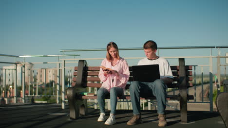 hombre y mujer sentados al aire libre estudiando juntos; niño escribiendo en la computadora portátil mientras mira a la señora leyendo de su libro, el fondo urbano presenta edificios de oficinas