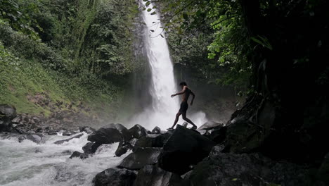 man climbs over rocks in riverbed with rushing waterfall behind, slow motion