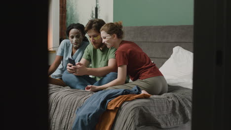 Drei-Mitbewohner-Mit-Gesichtsmaske-Schauen-Smartphone-Auf-Dem-Bett-In-Schlafzimmer-1