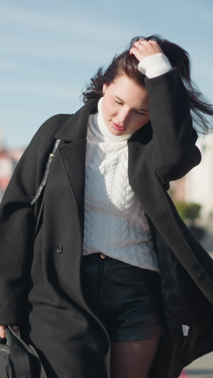 검은 코트, 짧은 가죽 바지,  스웨터를 입은 여인이 검은 드백을 들고 머리를 뒤로 조정하고, 도시 환경에서 태양이 그녀의 얼굴에서 반사됩니다.