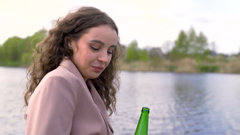 mujer morena rizada feliz hablando con su amiga y bebiendo cerveza en la orilla del río