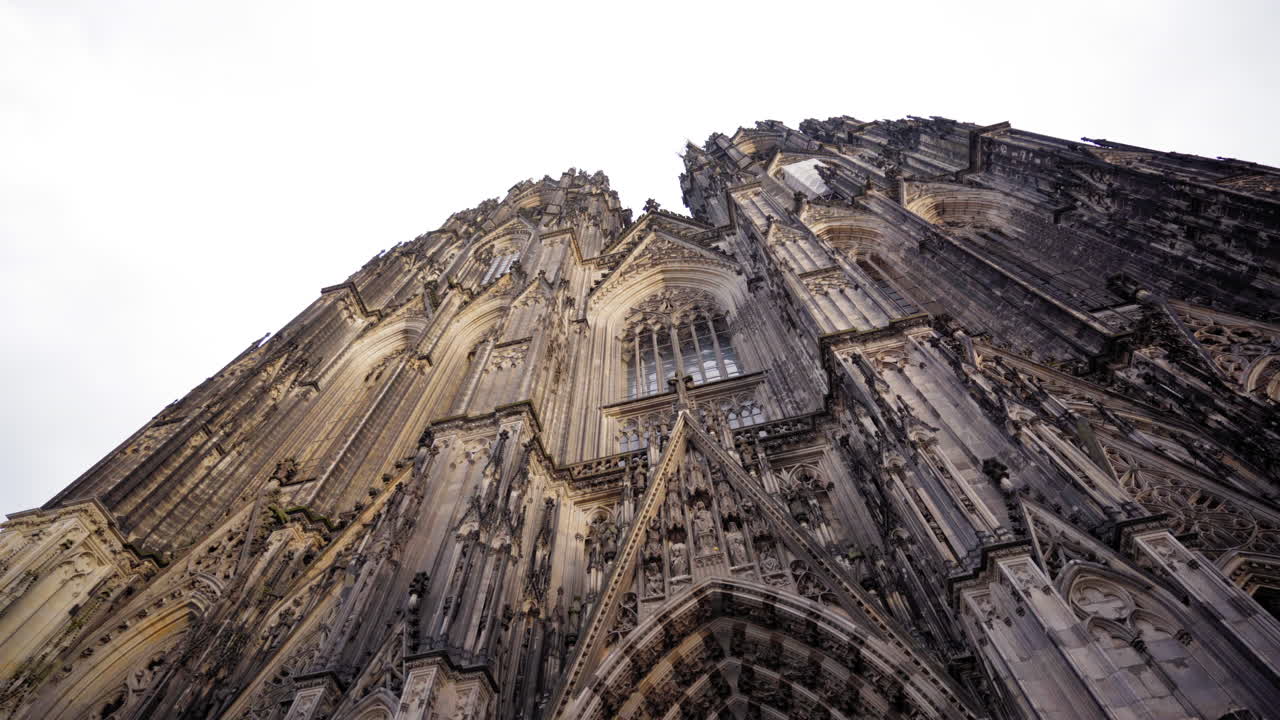 Upward view of the Cologne Cathedral's intricate Gothic facade with its stunning spires and detailed stone carvings, captured on a cloudy day in Cologne, Germany.