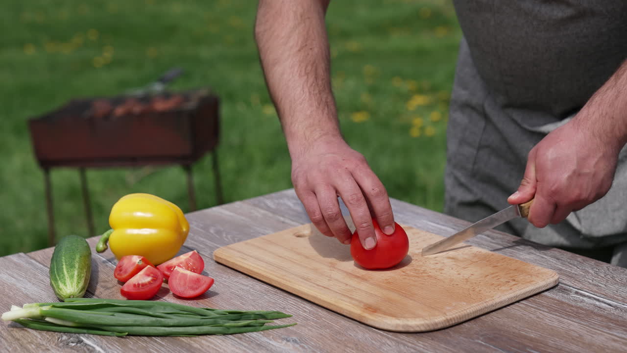 Man cutting tomato on a board outdoors. Preparing dinner at dacha. Man cuts vegetable into four parts on a table before the picnic.