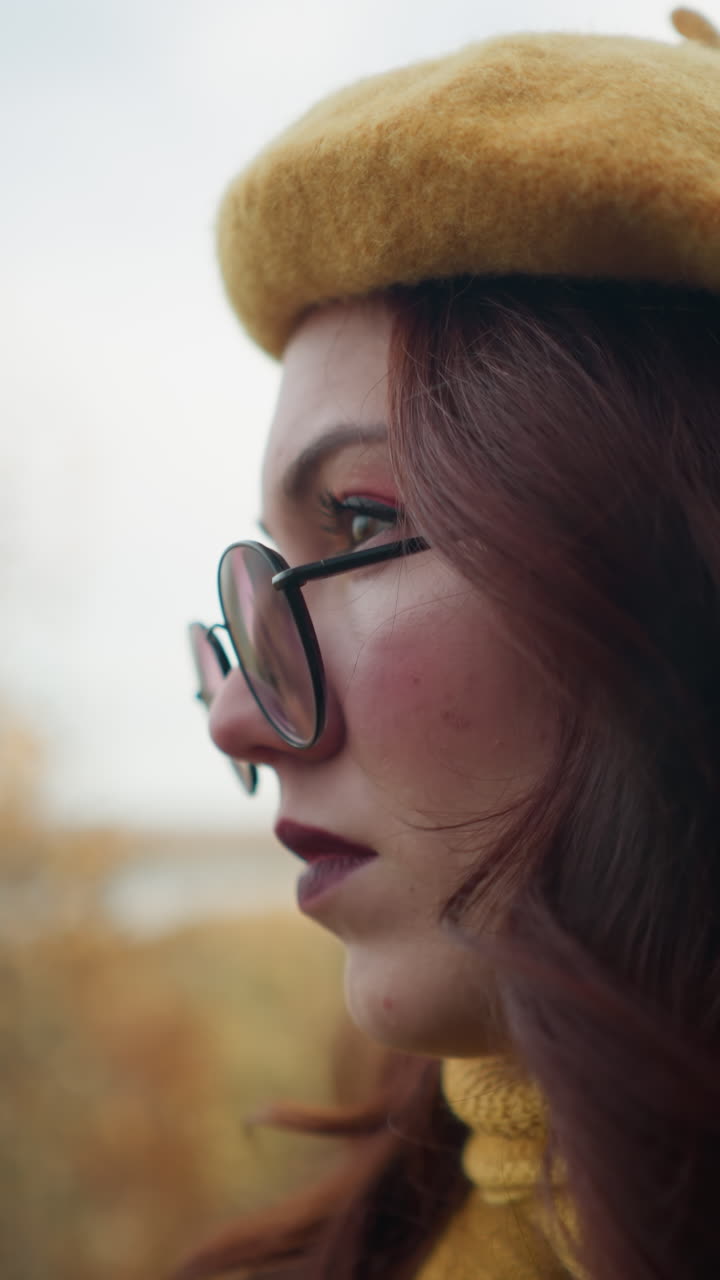Primo piano di una donna elegante con basco giallo e occhiali, che guarda in lontananza con un sorriso dolce; alberi autunnali e un fiume creano uno sfondo sfocato, aggiungendo calore e profondità all'espressione pensierosa.