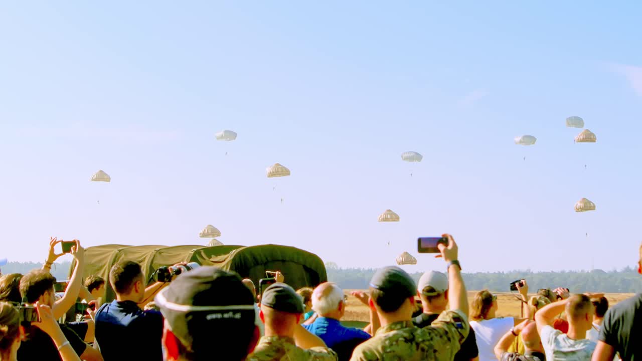 Spectators capture the scene as multiple parachutists descend with tan parachutes during a mass drop event at Airborne Ede, Ginkelse Heide. Crowd's excitement is evident as they watch the jumpers land