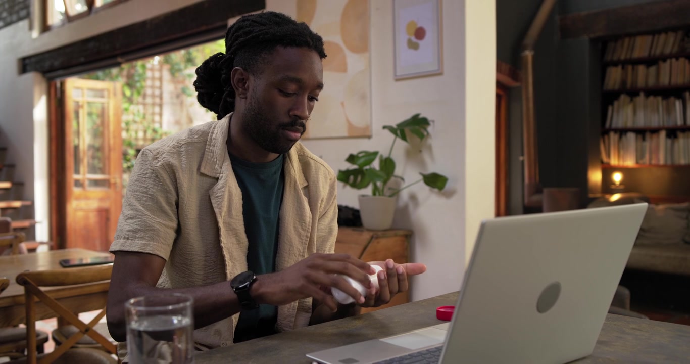 African male working on laptop at table checking bottle label, taking pills for relief with water