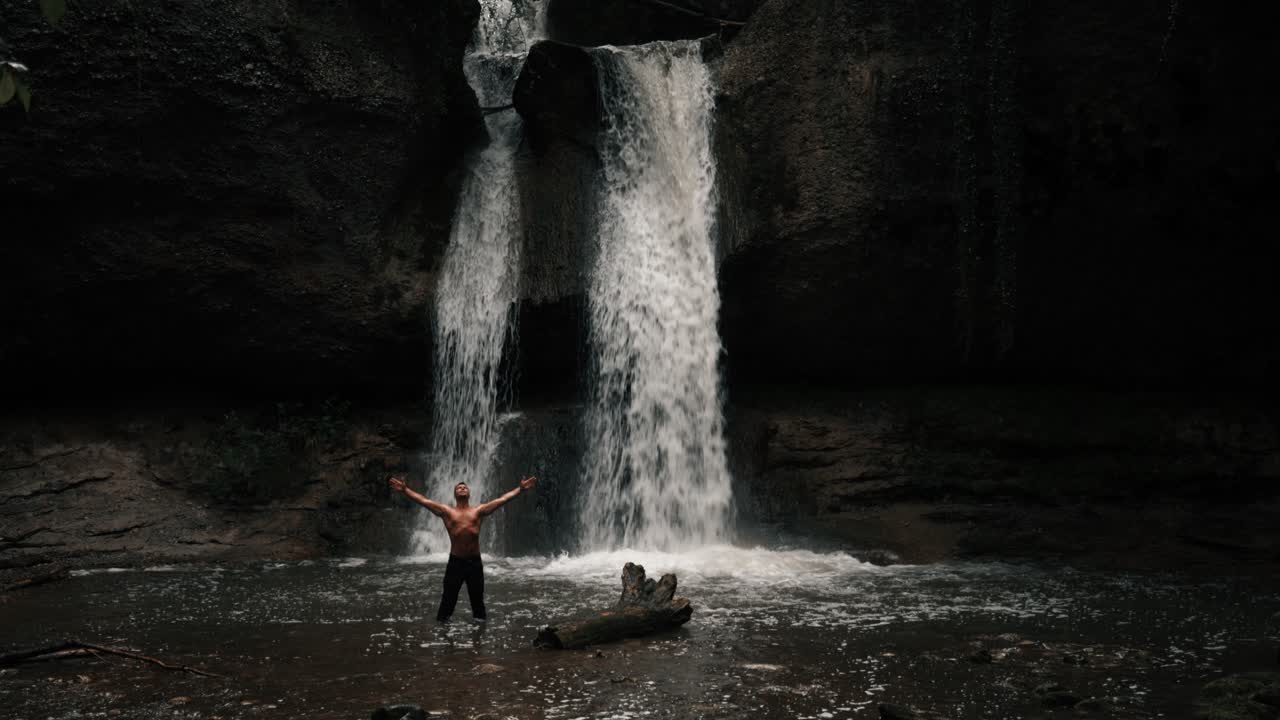 A man stands in the water in front of a waterfall in a mystical forest. It's raining and cloudy. He's shirtless and stretches his hands in the air. Beautiful, epic natural landscape. Slow motion shot.