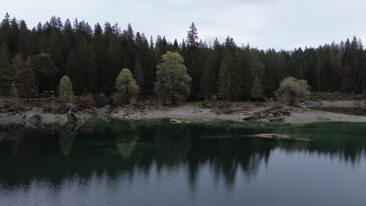 Movimento di allontanamento di un drone aereo che rivela un lago limpido color smeraldo circondato da una fitta foresta con riflessi sull'acqua calma, creando una scena di paesaggio naturale e pacifico