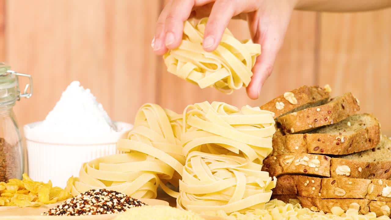 Hands arrange tagliatelle and bread on a wooden surface. Bright lighting highlights the rustic kitchen setting and food textures