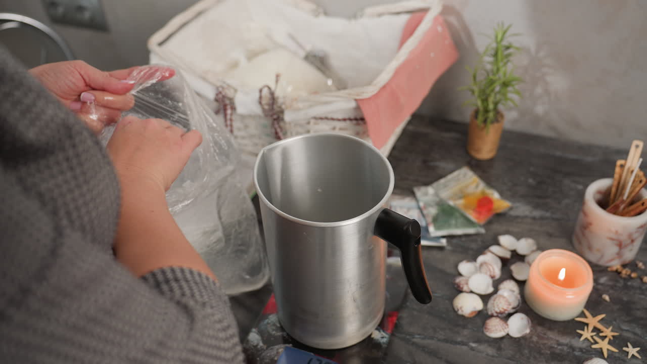 Woman holding clear ice from plastic bag and putting it inside metal jug on marble table as water splashes slightly, surrounded by shells, candle, art supplies, and decorative elements