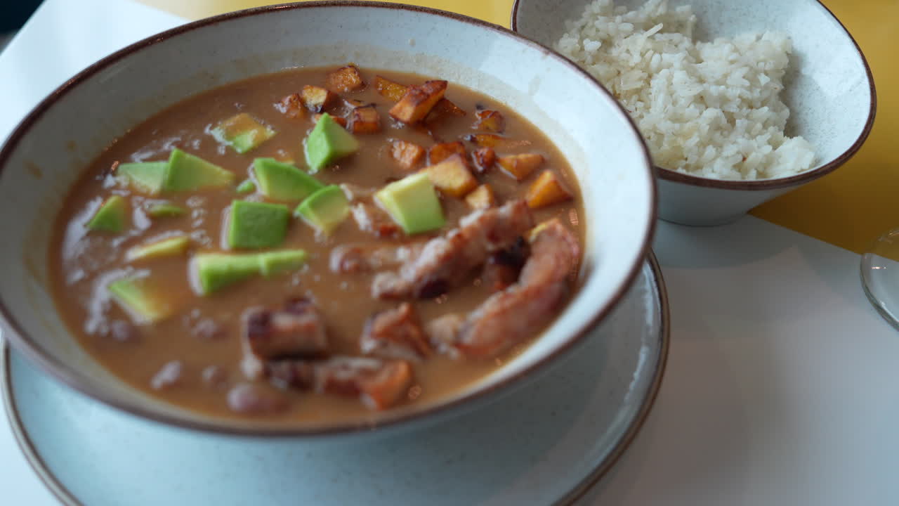 Uma visão detalhada da cazuela de frijoles colombiana com abacate por cima e servida com arroz. As cores e texturas vibrantes são capturadas neste close-up, perfeito para entusiastas da culinária