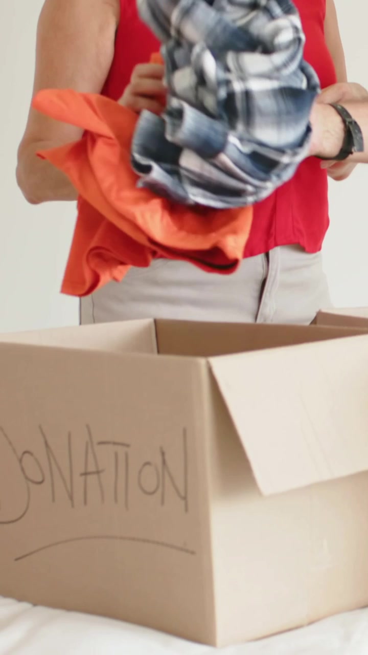 Vertical video: Picking orange shirt, woman in red blouse folding clothes into donation box on bed