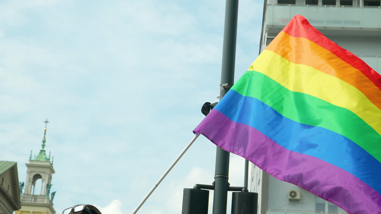 bandera lgbt unida a un largo poste blanco ondeando en el viento - en el fondo infraestructura urbana y cielo azul con nubes