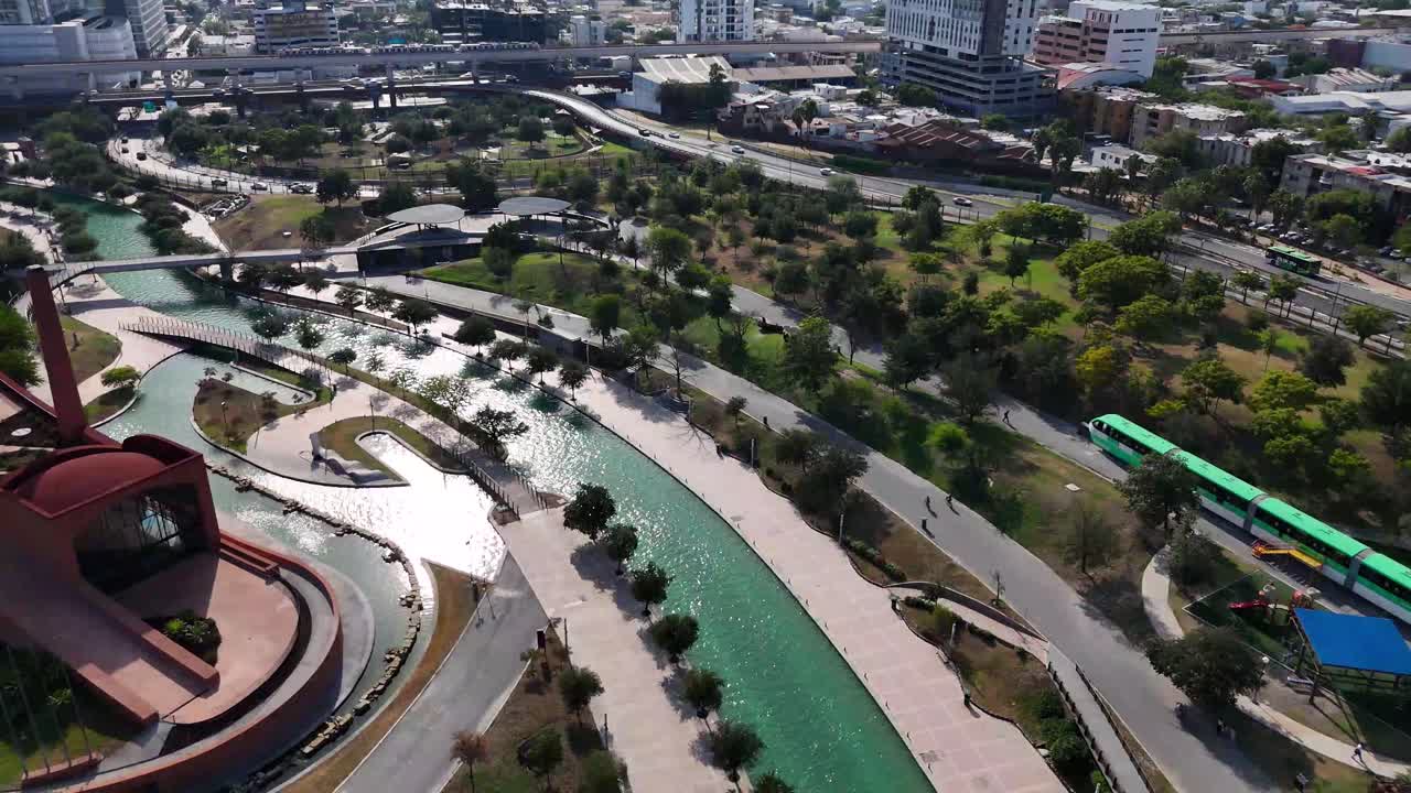 An artificial river in the middle of a dense city in Nuevo Leon, Mexico. In the background, you can see the subway and cars traveling through the streets on a sunny day, . 4K 60fps
