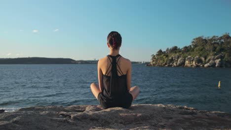 Wide-shot-of-young-female-seated-legs-crossed-by-the-water-meditating