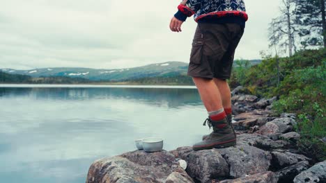 Hombre-En-La-Orilla-Rocosa-Del-Lago-Lavando-El-Tazón-De-Camping