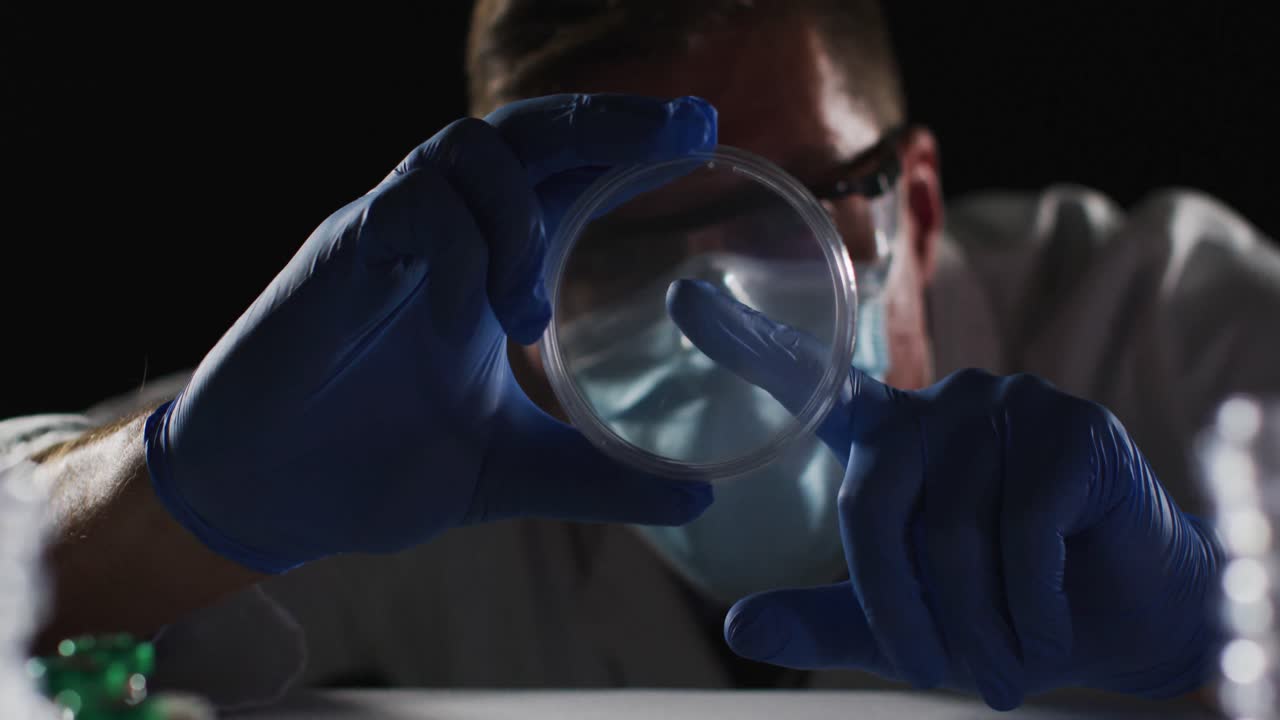 médico caucásico de sexo masculino con máscara facial y guantes inspeccionando la placa de petri
