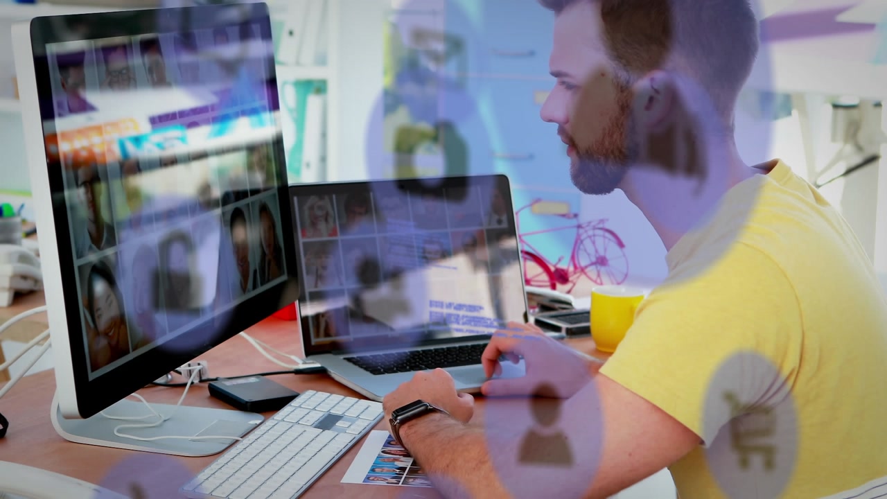 A male executive is looking at photos on a laptop and computer at a desk in an office
