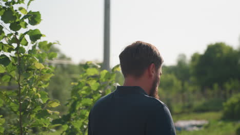 bearded man with rifle walks toward frame building in sunny yard, focused gaze while handling firearm, rural homestead background with garden and trees