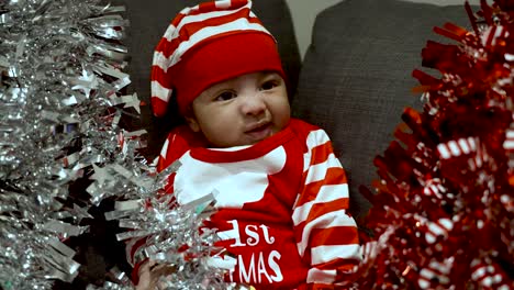 adorable 2 month old baby boy wearing red christmas outfit and hat and pulling on red and silver tinsel surrounded him on sofa