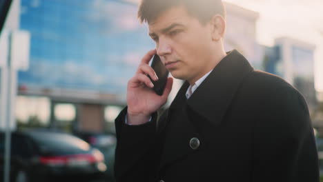 man in black coat adjusts phone under jaw while continuing call and nodding in agreement, background features modern glass buildings, parked cars, and warm sunlight