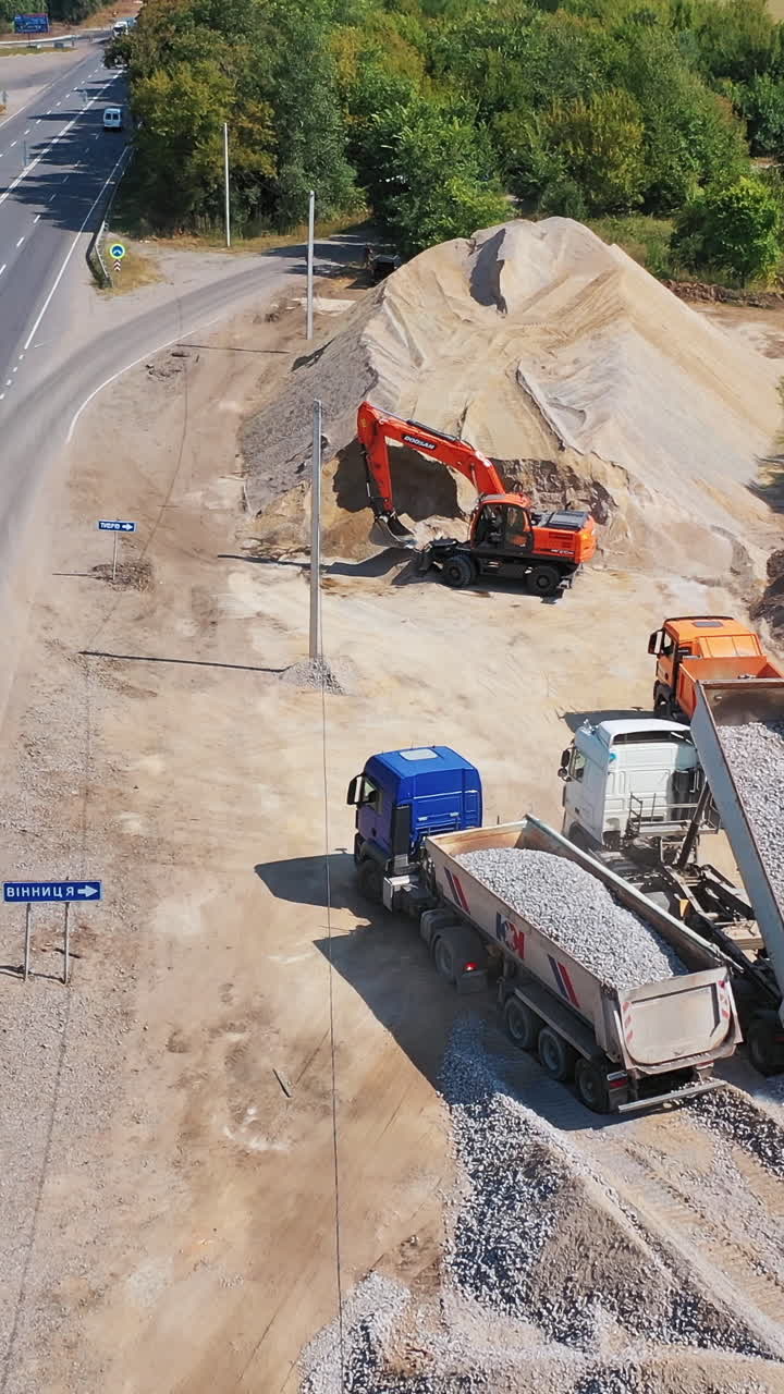 Trucks pouring out granite stone. Heavy machinery carry metal for construction new asphalt road near the highway. Aerial view. Vertical video