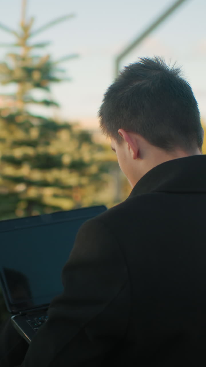 디자이너가 야외에 앉아 다리를 은 채 노트북을 작동하고 있습니다. 평화로운 환경에서 배경에는 움직이는 자동차, 근처에 주차된 자동차, 도시 구조물 및 초록색이 포함됩니다.