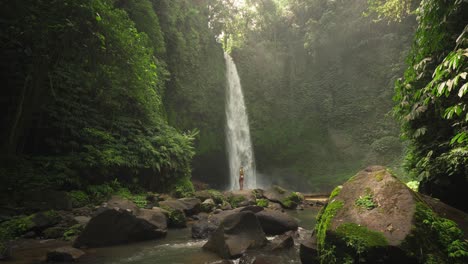 Magical-Nungnung-waterfall-with-beautiful-fit-blond-woman-posing-on-rock,-dreamy-sunlight