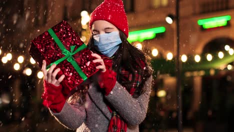 retrato de una niña caucásica feliz con bufanda sosteniendo un regalo y sonriendo a la cámara mientras nieva en la calle en navidad