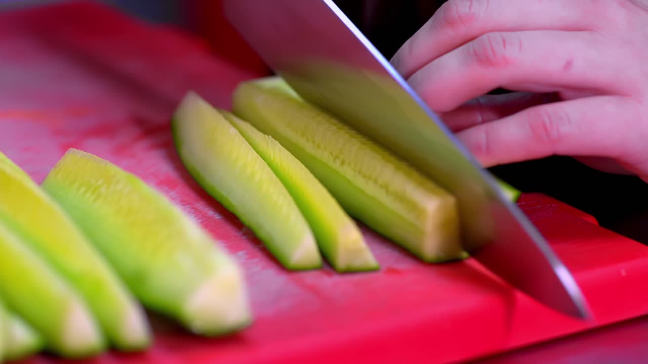 Hands slicing fresh cucumber on red cutting board with sharp knife in closeup