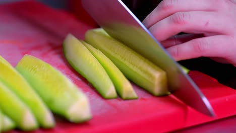 hands slicing fresh cucumber on red cutting board with sharp knife in closeup