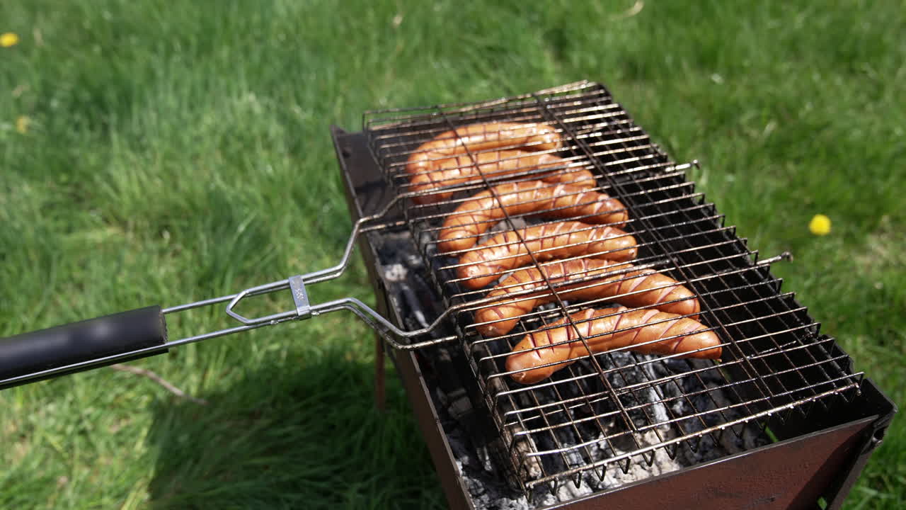 Roasting sausages on a grill outdoors. Delicious hot dog sausages grilling in smoke among green nature. Cooking meat sausages for a picnic.