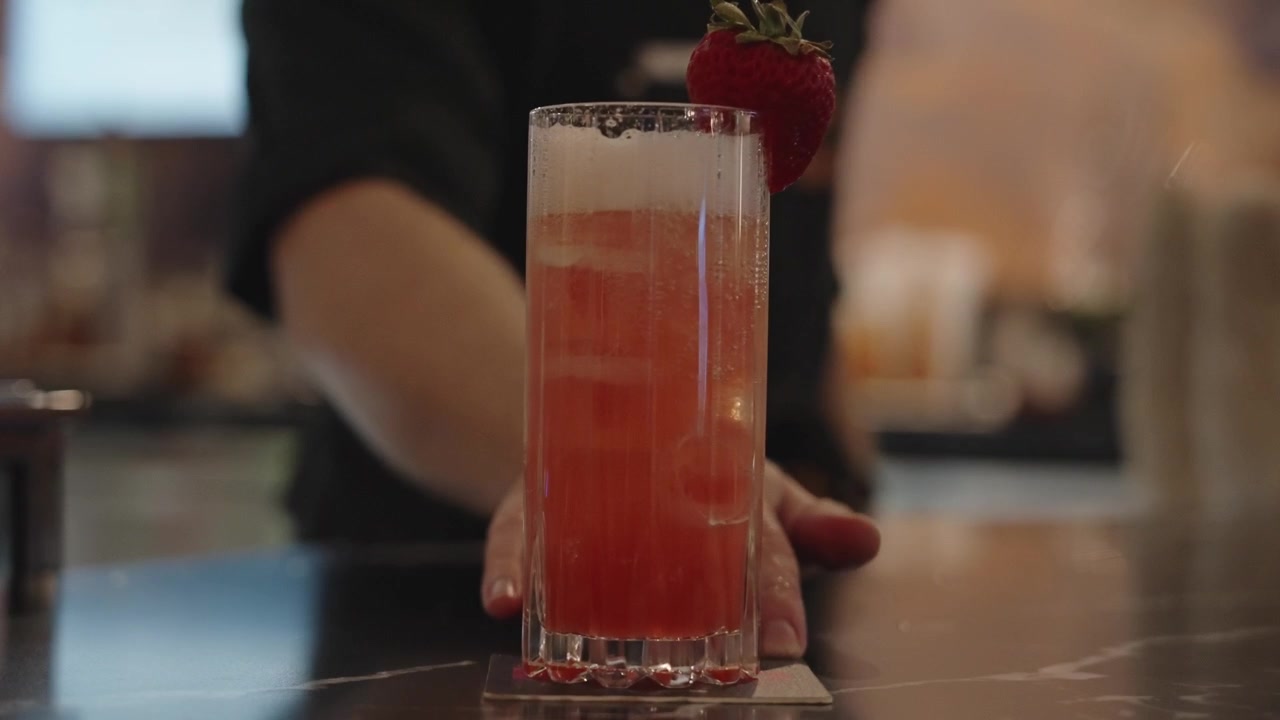 4K-optagelse af en bartender på et luksushotel, der skubber en iskold lyserød cocktail pyntet med et jordbær i et højt glas mod kameraet, hvilket fremhæver elegant barservice.