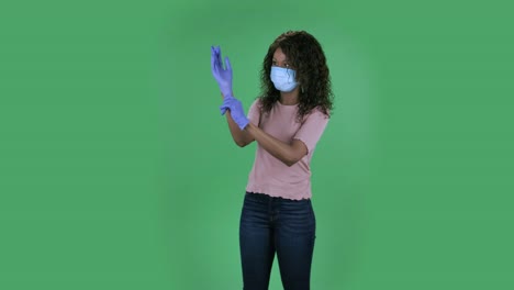 portrait of beautiful african american young woman in medical mask is looking at camera and puts on rubber gloves. burning brunette with wavy hair in jeans and a beige blouse on a green screen in the studio. health protection corona virus concept