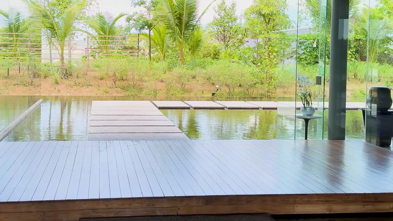 A serene view of a wooden deck extending over calm water, surrounded by lush greenery and palm trees.
