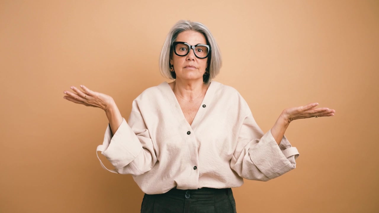 Mature Woman Shrugging Shoulders in Confusion Against Beige Background