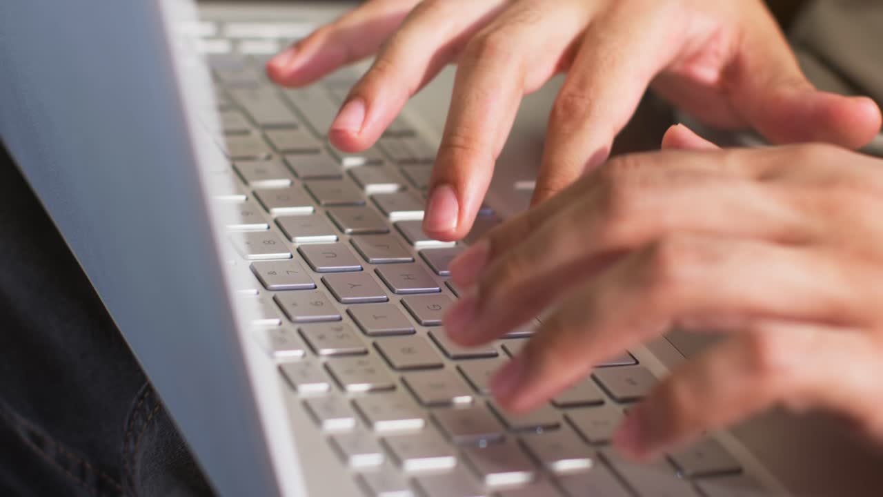 Mani maschili adulte che digitano muovendosi sulla tastiera del laptop appoggiato sulle ginocchia nella stanza, con maniche corte, laptop argentato.