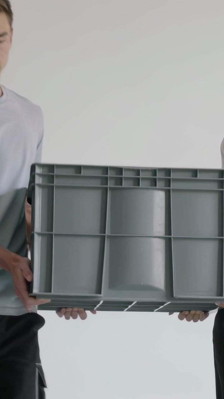 Vertical video: Grasping handles, two helpers carrying gray crate across white room, moving items