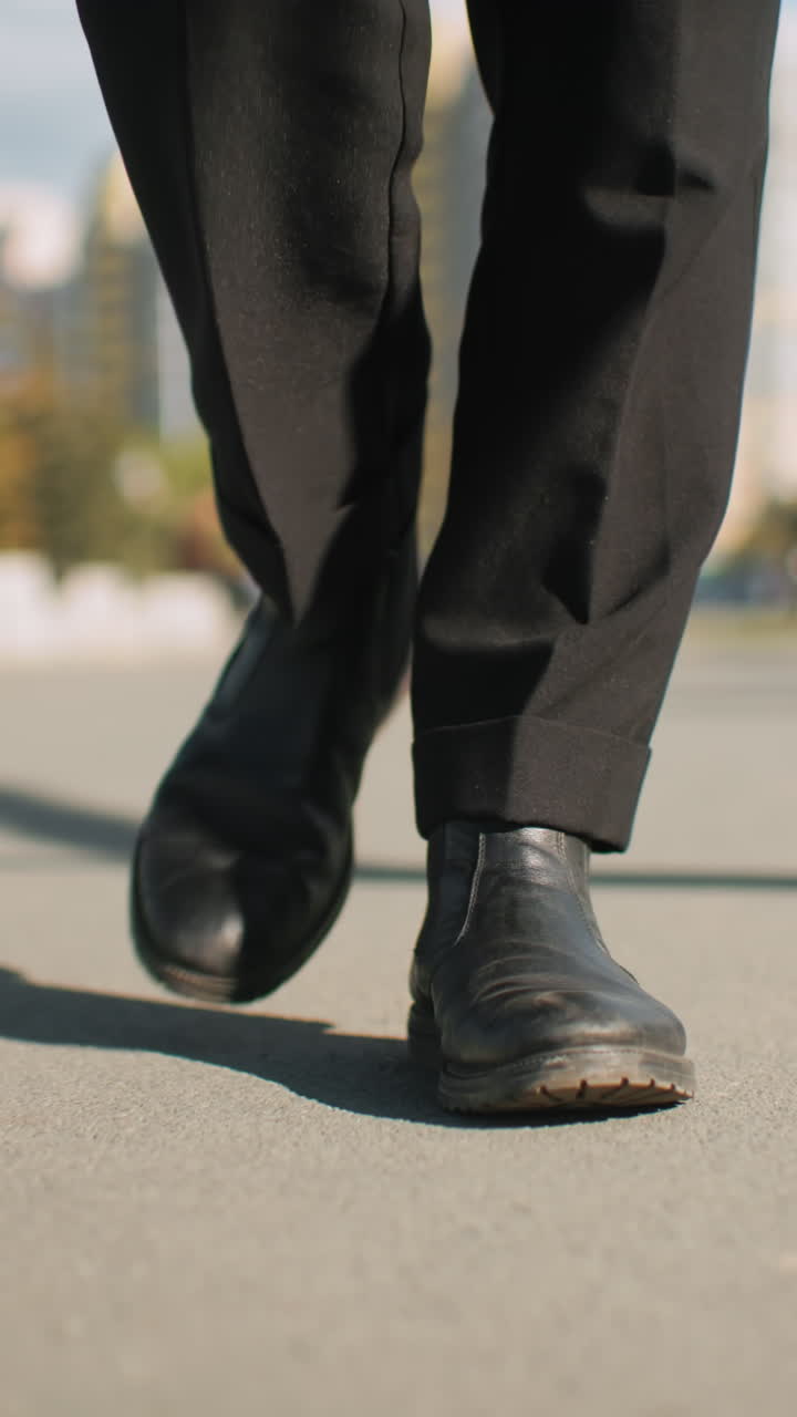 vista de la pierna de un individuo que lleva pantalones y zapatos negros caminando con confianza al aire libre en un camino asfalto, la sombra proyectada en el suelo, rodeado de árboles y fondo urbano